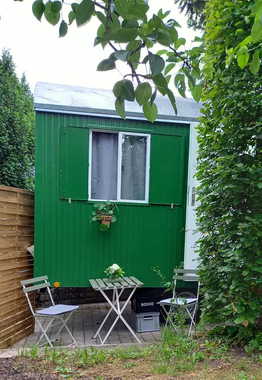 Grüner Bauwagen mit Gardine am Fenster, davor Bistrotisch und Stühle